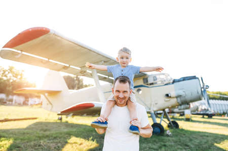 A dad and toddler spend time outdoors in an airplanes museum. A young father carries baby boy on his shoulders, a son spread his arms like wings of an airplanesの写真素材