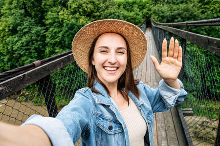 Always in touch. Video screen of girl in hat and casual jeans jacket takes a selfie on suspension cable bridge surrounded foggy forest. A cheerful woman holds a phone and looks at camera.の写真素材