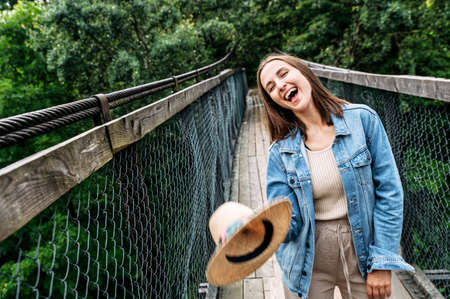 Young cheerful woman stands on suspension cable bridge among wild forest, throws her straw hat and laughing happily. Good mood for adventuresの写真素材