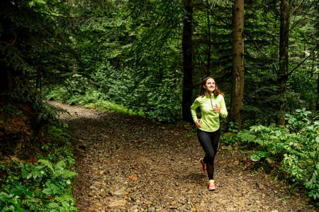 A girl in neon green long sleeve shirt is running on the forest path. Morning jog outdoors