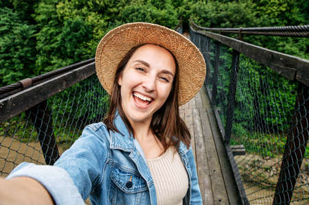 A girl in straw hat stands on the suspension bridge, holds a smartphone in hand and takes a selfie with a cheerful smileの写真素材