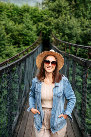 Traveler girl in hat and casual jeans jacket stands on suspension cable bridge surrounded foggy forestの写真素材