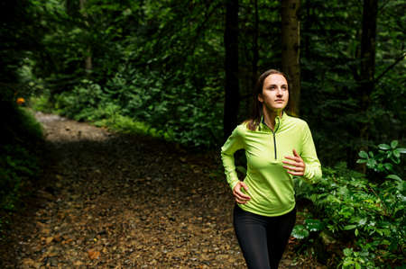 A woman in sports long sleeve shirt is running on dirt road through forest. Healthy lifestyle...