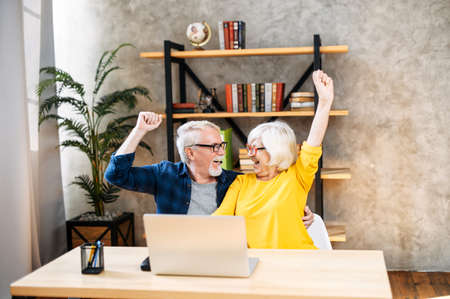 Overjoyed mature grey-haired husband and wife looks on laptop indoor. Excited senior couple rais hands up in triumph reading good news on computer, winner conceptの写真素材