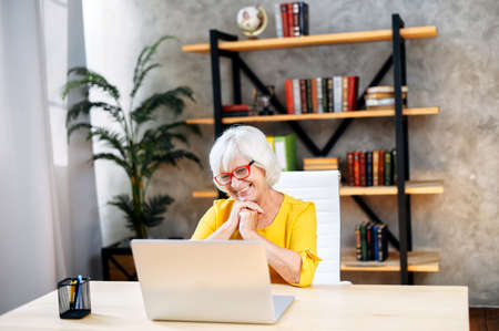 A touched gray-haired business lady in eyeglasses is looking at the laptop screen while sitting in a smart officeの写真素材