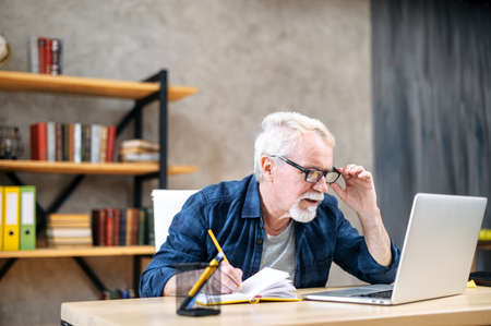 Focused older man in smart casual wear is using laptop computer for watching online classes and webinars, he is takes a notes at notebook, while sitting at contemporary home officeの写真素材