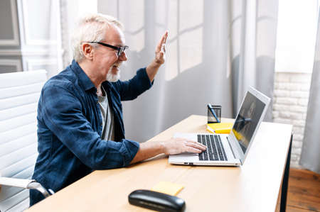A handsome mature man talking via video call on laptop while sitting at contemporary office. A senior man is waving at webcam cheerfullyの写真素材