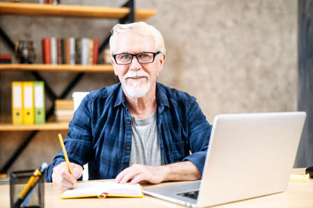 Handsome senior man in smart casual wear is using laptop computer for watching online classes and webinars, he is takes a notes at notebook, while sitting at contemporary home officeの写真素材