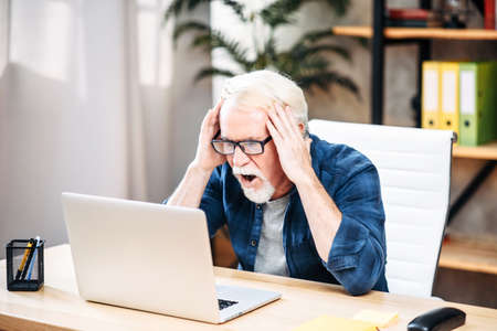 Senior man looks at the laptop screen with shock and surprise, he holding head in his handsの写真素材