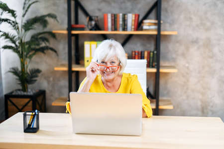 A beautiful senior business lady lowered her eyeglasses and looks at the camera while using laptop at home officeの写真素材