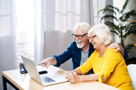 A cheerful elderly couple is web surfing the laptop indoors, a grey haired man points on the screen, a senior lady laughsの写真素材