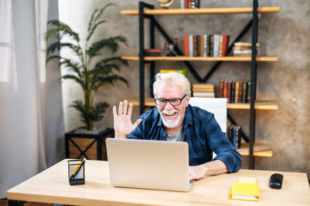 Handsome smiling older man with mustache and beard is using a laptop computer for online video call at home office. A gray-haired man is waving at webcamの写真素材