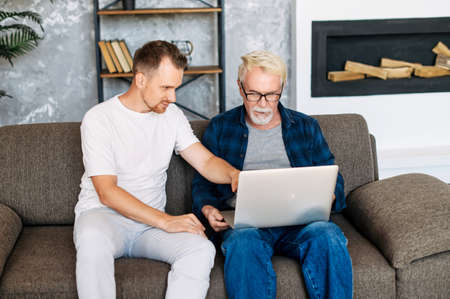Young adult son teaches old senior father using laptop sit on sofa, two men diverse generation spend time online with computer together at homeの写真素材