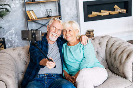 Modern grandparent are taking selfie by a smartphone on the selfie stick sitting at sofa in cozy living roomの写真素材