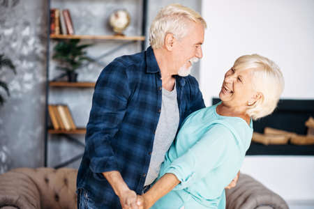 Portrait of modern romantic elderly couple is hugging, embracing gently, dancing at home. Senior spouses stand holding hands, looks to each other and laugh happilyの写真素材