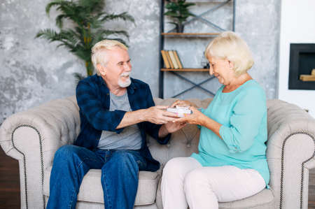 Elderly husband made a surprise for his wife. Senior couple sits on the sofa at home, a gray haired man is presenting gift to elderly pleasantly surprised womanの写真素材