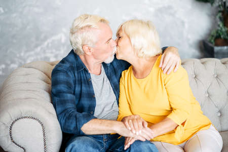 Elderly gray-haired couple in love is kissing. Senior spouses sit on cozy sofa at home in embrasesの写真素材