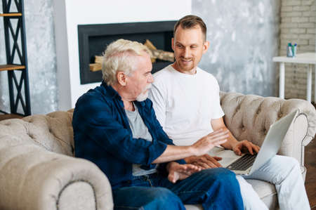 Cheerful two generation men family old senior father and smiling young adult son talking sitting on the comfortable sofa at home, using laptop for websurfing, online shoppingの写真素材