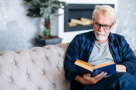 Senior man spends leisure time reading book sitting on the comfortable couch at cozy home. A gray-haired older man in eyeglasses immersed in readingの写真素材