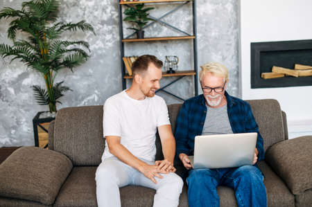 Young man teaching gray-haired senior man to using computer. Two multigeneration men sit on the sofa with a laptopの写真素材