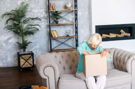 An elderly woman unpacks a shipping box with interest. A gray-haired elderly woman sits in the living room and looks into a cardboard box with online orderの写真素材