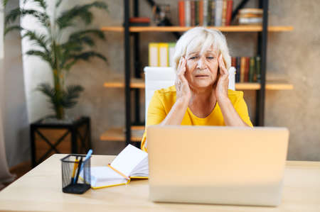 Tired senior business woman is using laptop for work indoor. A beautiful older lady has headache...