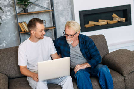 Young man teaching gray-haired senior man to using computer. Two multigeneration men sit on the sofa with a laptopの写真素材