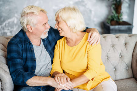 Elderly couple looks to each other eyes with love and care spending leisure at home. A bearded senior husband and gray-haired beautiful wife holding hands sitting on the sofaの写真素材
