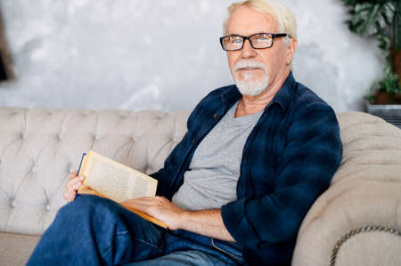 Mature man loves reading in free time. A gray-haired older man in eyeglasses sits on the sofa with a book in hands and looks into a camera with a serious faceの写真素材