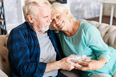 Elderly couple has anniversary or celebrates some important date. Senior gallant man is giving a gift to charming smiling elderly womanの写真素材