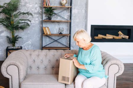 Elderly lady got delivery of online order. Senior woman is unpacking cardboard shipping box sitting...