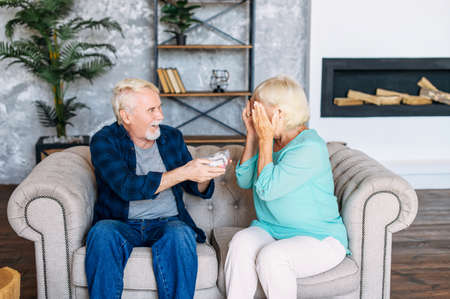A romantic senior couple at home. Mature husband is giving a gift in a cute box to his wife, she is waiting with eyes closed. Surprise to anniversaryの写真素材