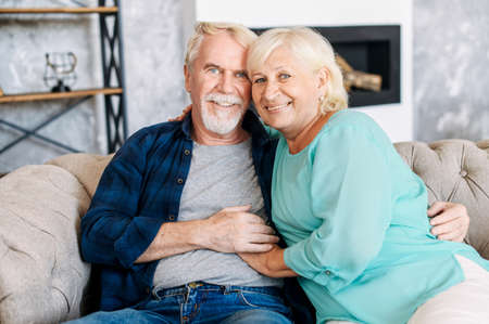 Senior happy couple is holding hands and hugging sitting together on comfortable sofa at home. Concept of support and caring in marriageの写真素材