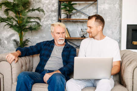 Young adult son teaches old senior father using laptop sit on sofa, two men diverse generation spend time online with computer together at homeの写真素材