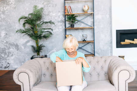 Elderly lady got delivery of online order. Senior woman is unpacking cardboard shipping box sitting on the sofa in cozy living roomの写真素材