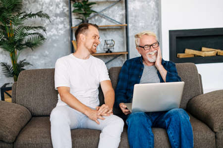 Young man teaching gray-haired senior man to using computer. Two multigeneration men sit on the sofa with a laptopの写真素材