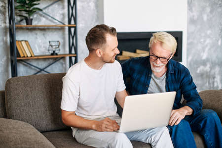 Young adult son teaches old senior father using laptop sit on sofa, two men diverse generation spend time online with computer together at homeの写真素材