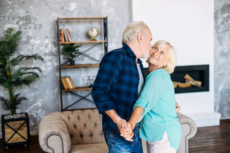 Happy senior wife and husband hugging at home, a man is kissing woman. Romantic middle-age couple has fun, dancing and laughingの写真素材