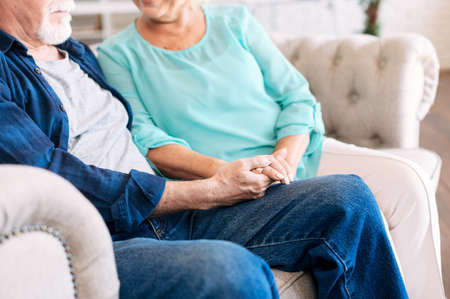 Senior couple holding hands sitting together at home, faces is not visible. Concept of support and caring in marriageの写真素材