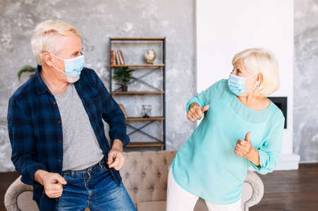Positive and cheerful senior couple wearing medicine masks has fun together at home during quarantine. Elderly spouses dancing with surgery masks on their facesの写真素材