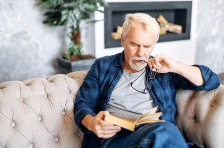 Mature man loves reading in free time. A thoughtful gray-haired older man sits on the sofa with a book in hands in a cozy living roomの写真素材
