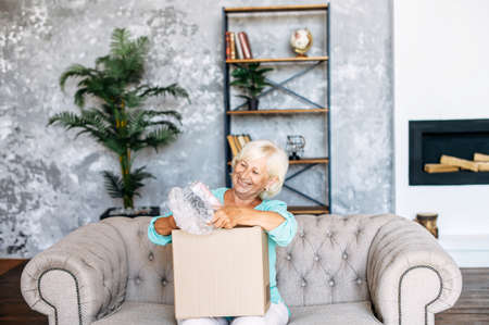 An elderly woman unpacks a shipping box with joy and interest. Smiling gray-haired woman sits in the...