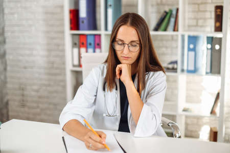 Focused young female doctor with a stethoscope on the shoulders sits at the desk and fills out an application form at office in clinic while has video call with a patient. Virtual appointment conceptの写真素材