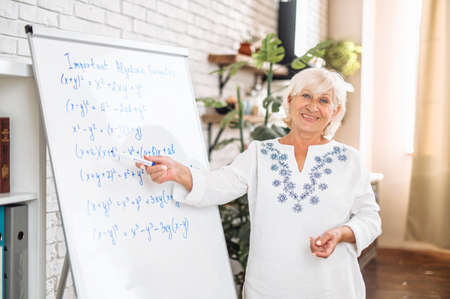 A senior woman in smart casual shirt is writing on flipchart and smiles. Online teacher, tutor. Side viewの写真素材