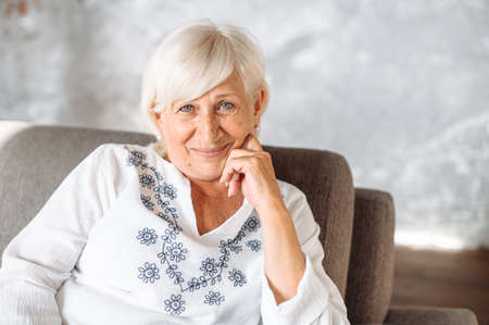Portrait of charming senior woman indoors. Gray haired elderly lady sits on the comfortable sofa at home, looks at camera and smilesの写真素材