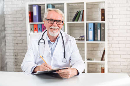 Smiling senior doctor in eyeglasses sits in medical office. A mature MD in white uniform with a stethoscope looks at the camera and writes a report. Virtual visit concept, video callの写真素材