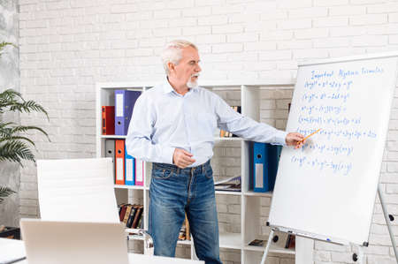 Confident senior male teacher conducts online classes in contemporary classroom. Older math professor explains something to online audience on laptop screen. Distance studying conceptの写真素材