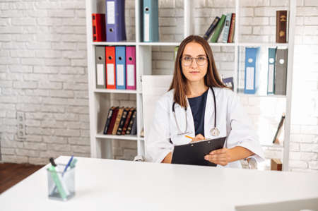 Focused young female doctor with a stethoscope on the shoulders sits at the desk and fills out an application form at office in clinic while has video call with a patient. Virtual appointment conceptの写真素材