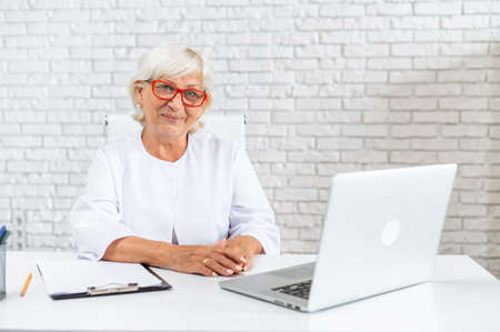 Mature gray haired female doctor is using laptop in hospital office. Senior woman in white formal coat on the white backgroundの写真素材