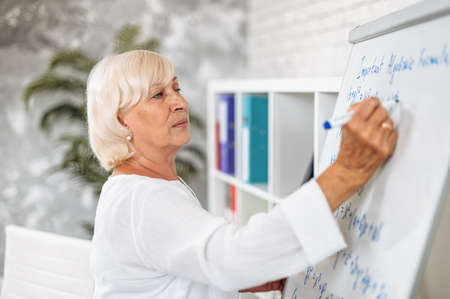 Concentrated senior female teacher writes algebra formulas on a whiteboard in modern classroomの写真素材